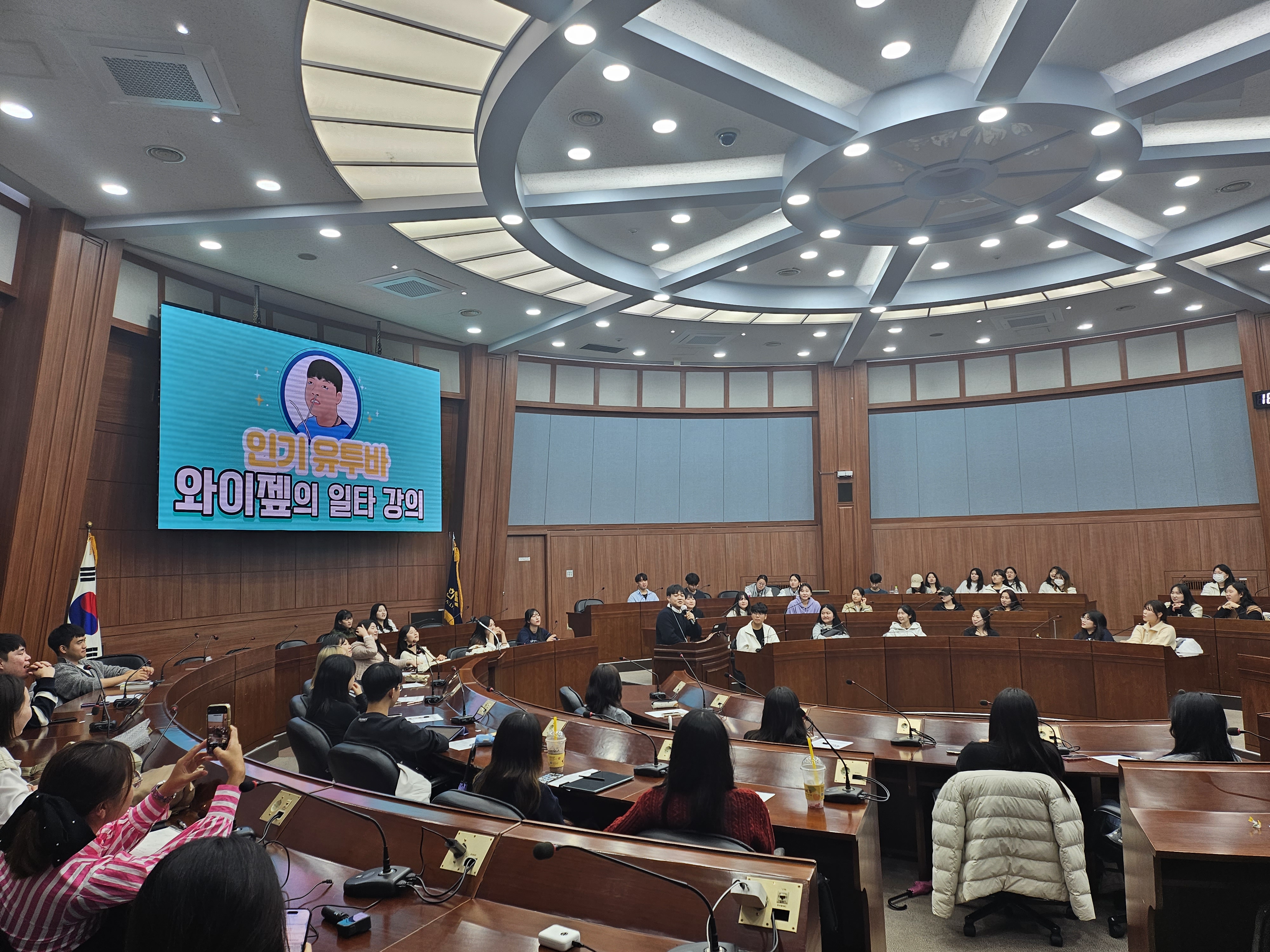 유튜버 와이젭이 원형 강의실에서 특강을 진행하고 있다. 한림대학교 간호대학 학생들이 주위에 앉아 강의를 듣고 있다. 강의실 벽면 화면에는 인기 유투바 와이젭의 일타 강의라고 적혀있다.