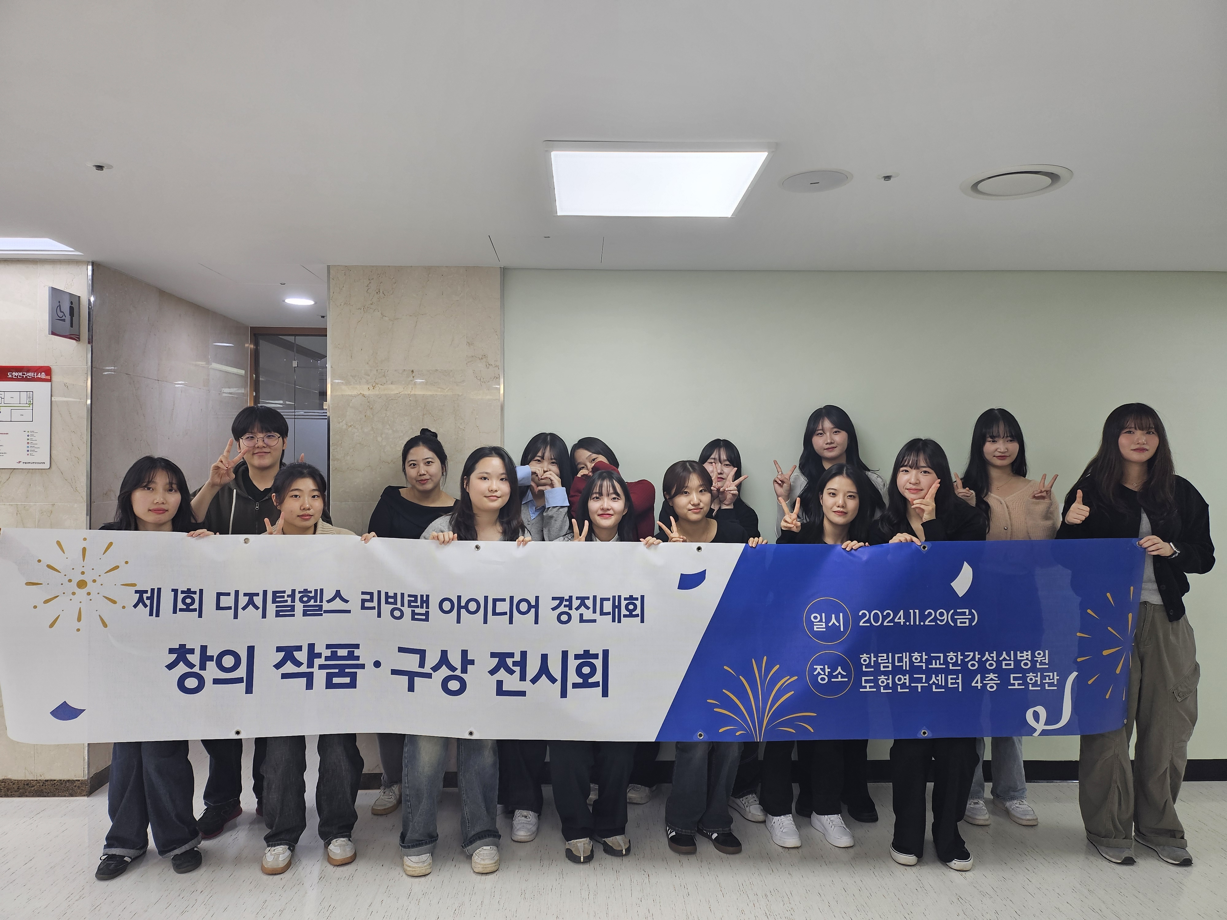 제 1회 디지털헬스 리빙랩 아이디어 경진대회 창의 작품 구상 전시회 현수막을 들고 간호대학 재학생들이 단체 사진을 찍고 있다.